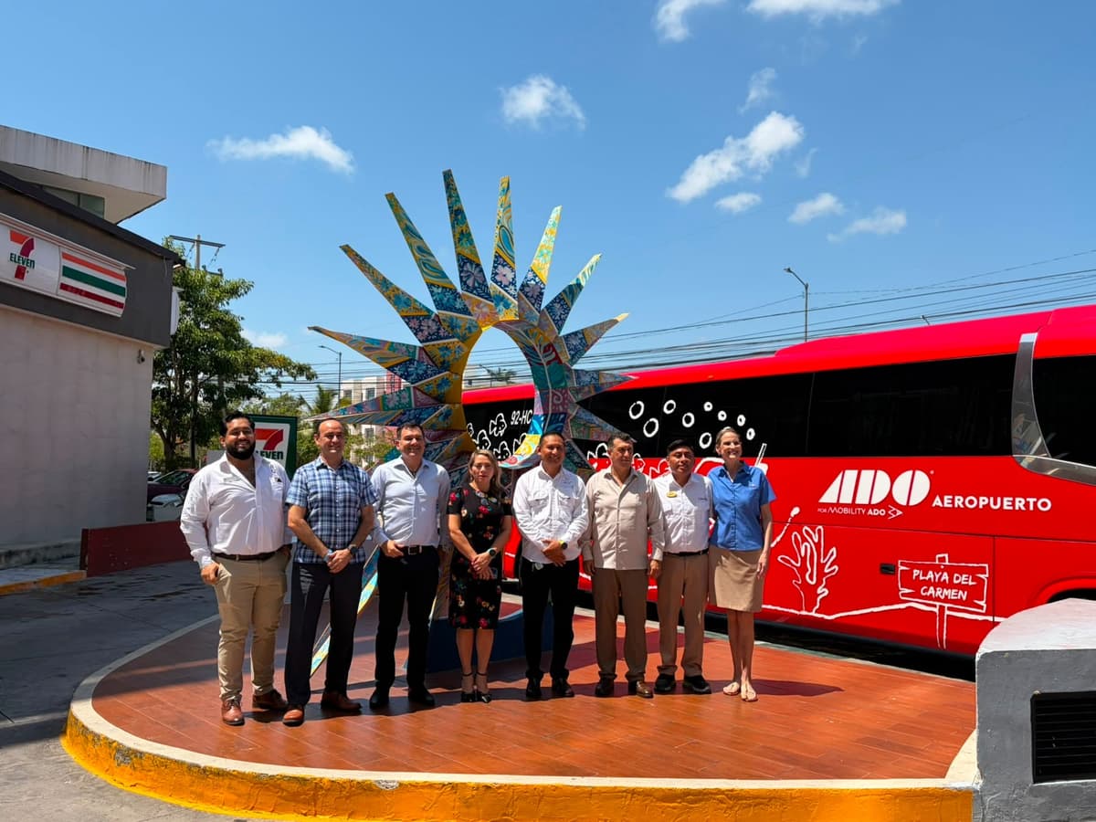 Grupo de personas posando frente a un autobús rojo de ADO Aeropuerto con la escultura de una mano decorada de fondo en Cancún.