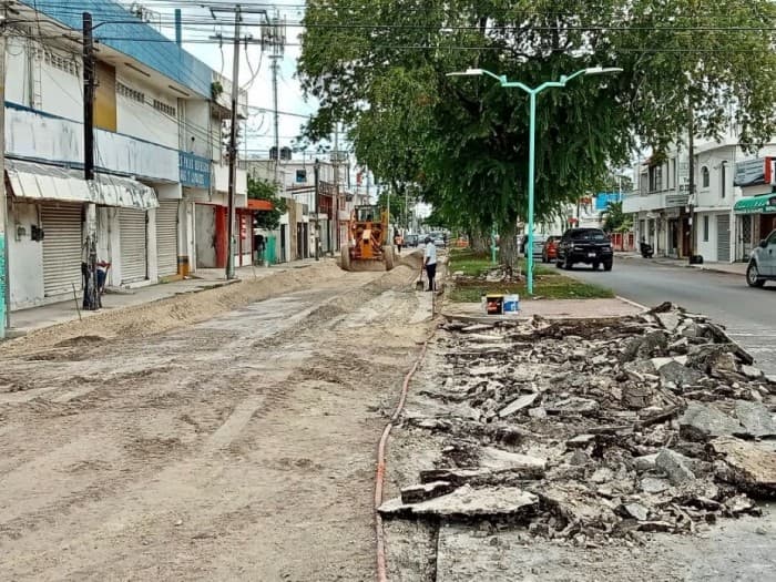 Calle en construcción con escombros y materiales de obra, edificios comerciales y residenciales a los lados, señalización vial verde.