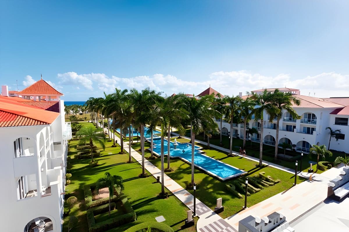 Vista aérea del complejo resort con piscinas, áreas verdes, palmeras y edificios blancos con techos de color naranja frente al mar.
