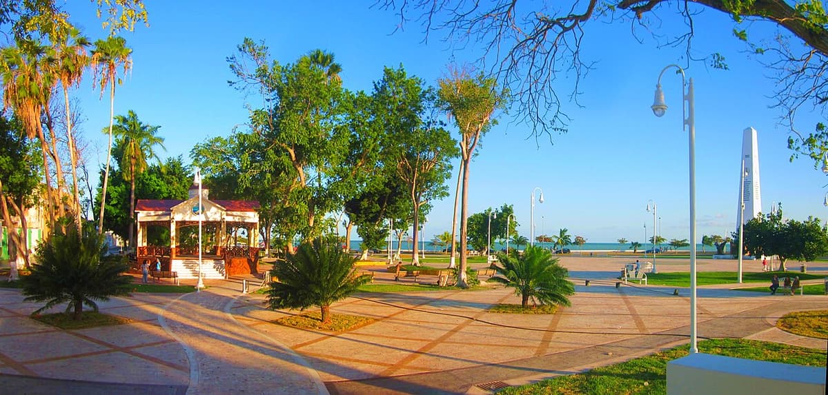 Parque turístico frente al mar en Quintana Roo con kiosco, palmeras y paseo peatonal con vista al agua en día soleado.